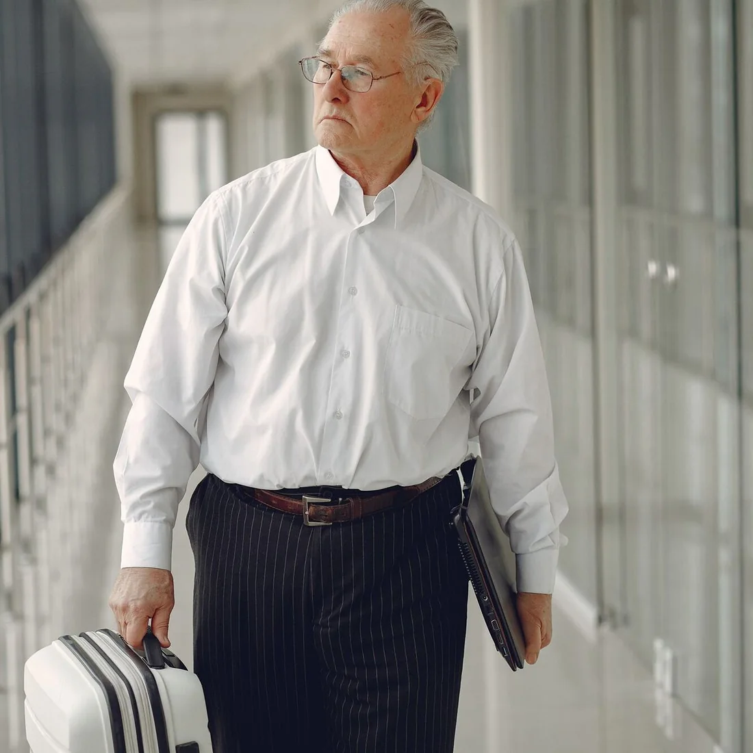 A man in a white shirt walks along a bright airport corridor with a suitcase and a laptop.