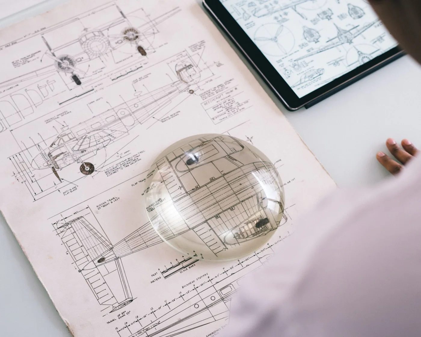 An aircraft engineering drawing under a magnifying glass, with a tablet showing the same aircraft beside it.